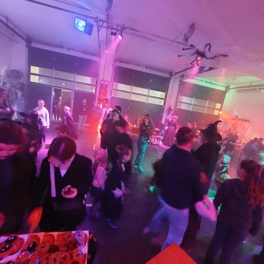 Ein Saal voller Menschen in Kostümen, einige in schicken Outfits, die herumlaufen und sich unterhalten. Der Ort ist schwach beleuchtet mit lila und grünen Lichtern. Ein Tisch mit Keksen und Donuts ist vorne aufgestellt.