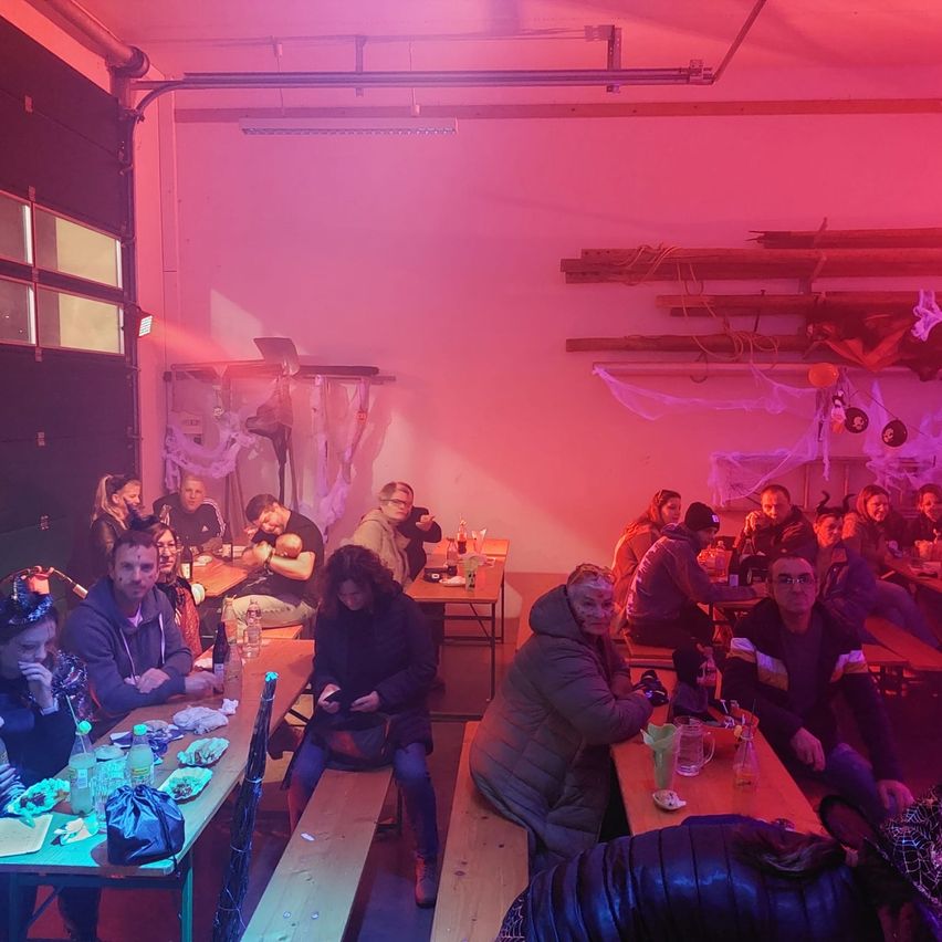 Eine Gruppe von Menschen sitzt an Tischen in einem Raum mit rosa Beleuchtung. Sie unterhalten sich und einige haben Essen auf ihren Tischen. Der Raum hat Holzwände und Regale mit hängenden Dekorationen.