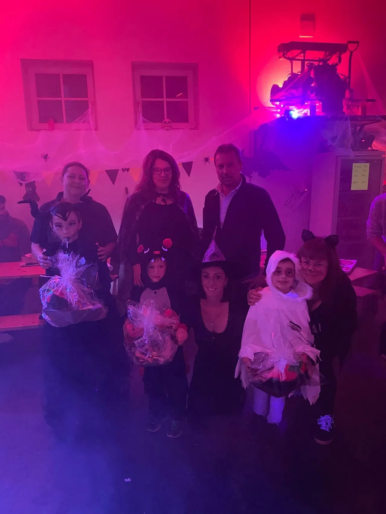 Eine Familie posiert für ein Foto auf einer Halloween-Party. Sie tragen Kostüme und halten Geschenktüten. Der Raum ist mit lila Lichtern und Halloween-Dekorationen geschmückt.