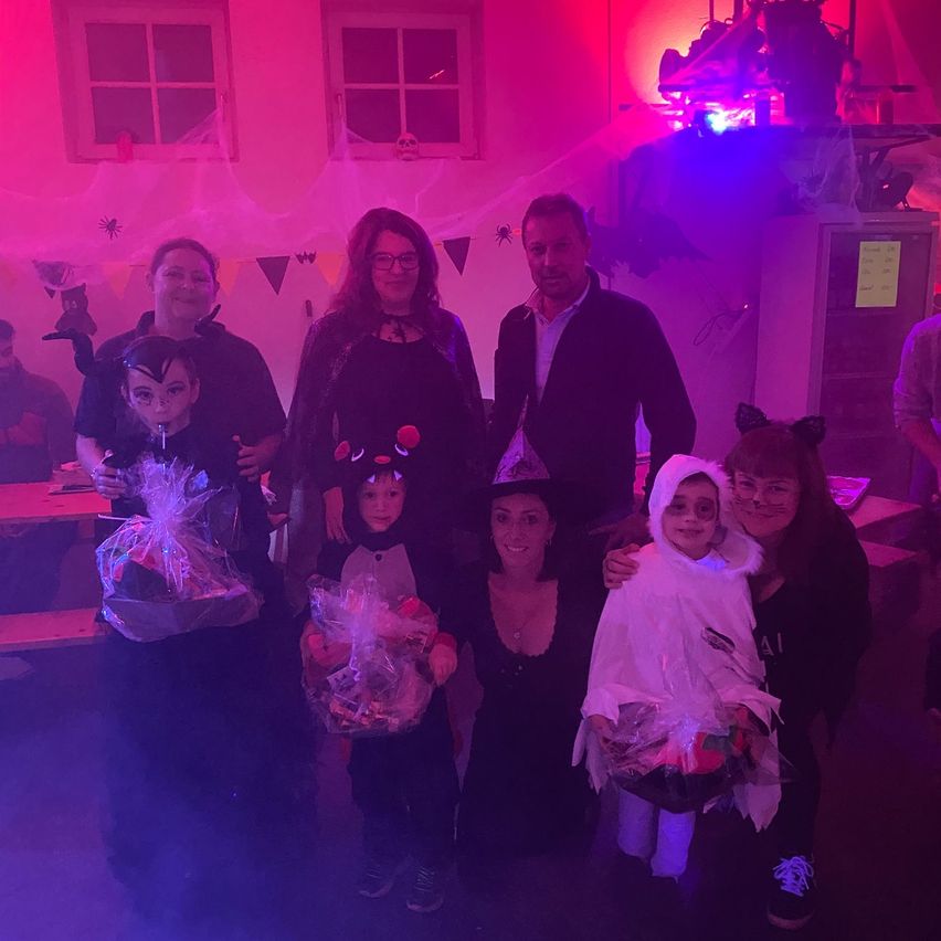Eine Familie posiert für ein Foto auf einer Halloween-Party. Sie tragen Kostüme und halten Geschenktüten. Der Raum ist mit lila Lichtern und Halloween-Dekorationen geschmückt.