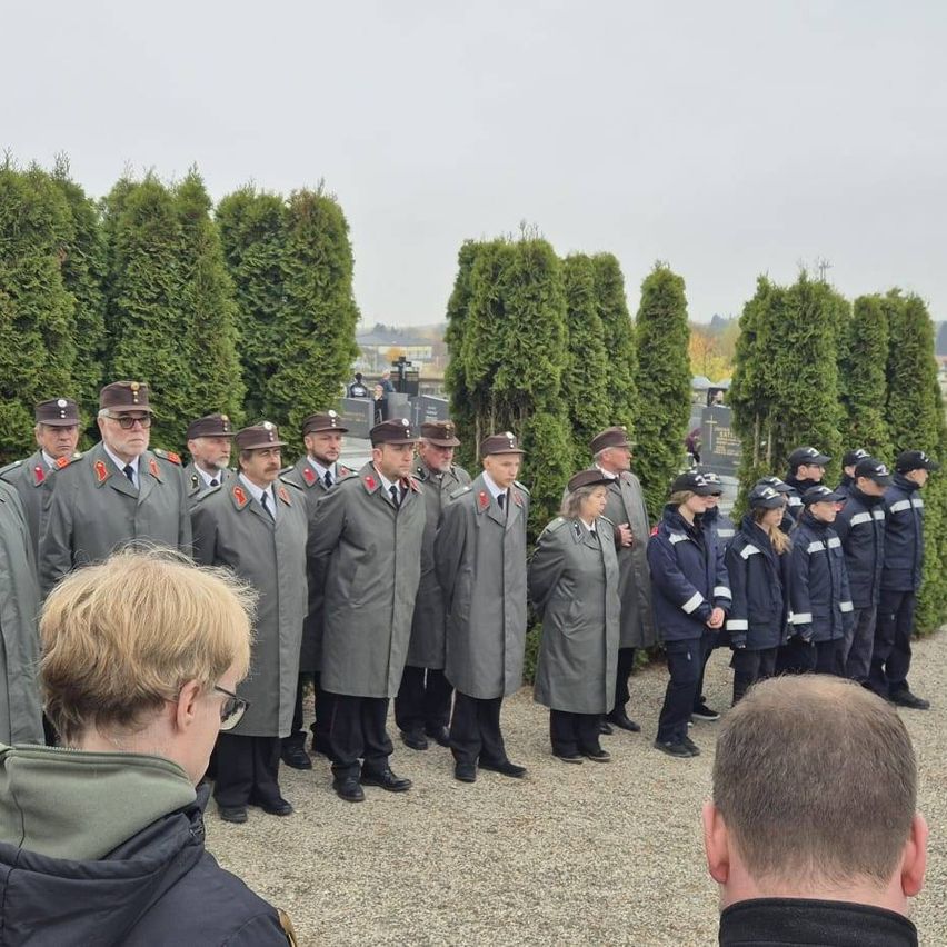 Eine Gruppe uniformierter Personen steht auf einem Friedhof, wobei eine Person mit Brille den Kameras den Rücken zukehrt.