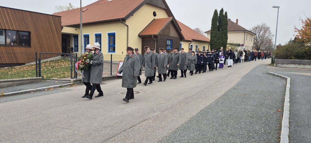 Eine formelle Prozession uniformierter Personen, einige mit Blumen, geht in einer Reihe auf einer Straße, hinter ihnen ein Gebäude.