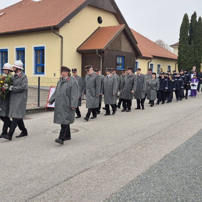 Eine formelle Prozession uniformierter Personen, einige mit Blumen, geht in einer Reihe auf einer Straße, hinter ihnen ein Gebäude.