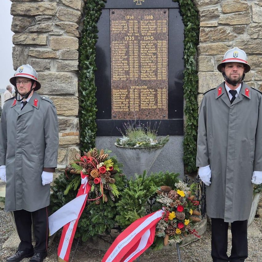 Zwei Männer in Uniform stehen neben einer Gedenktafel, umgeben von Kränzen und immergrünen Pflanzen. Die Tafel listet Namen und Jahre auf, möglicherweise zur Erinnerung an gefallene Soldaten.