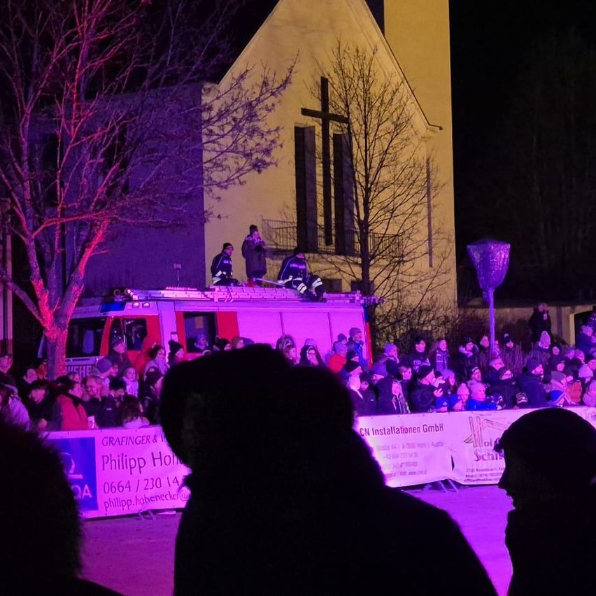Eine Menschenmenge versammelt sich vor einer Kirche, die mit lila Lichtern beleuchtet ist. Menschen stehen auf dem Dach eines Feuerwehrwagens und auf dem Balkon. Ein Banner zeigt eine Telefonnummer an.