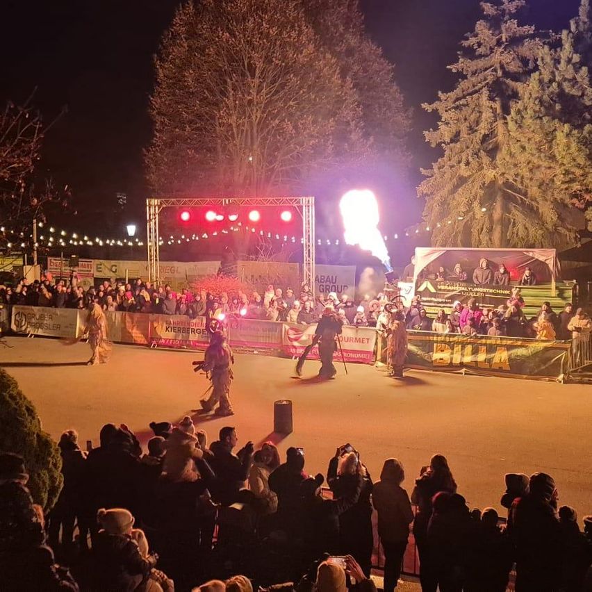 Ein nächtliches Event mit einer Menge, die Künstler auf einer Bühne mit roten Lichtern und einer großen Leinwand im Hintergrund beobachtet.