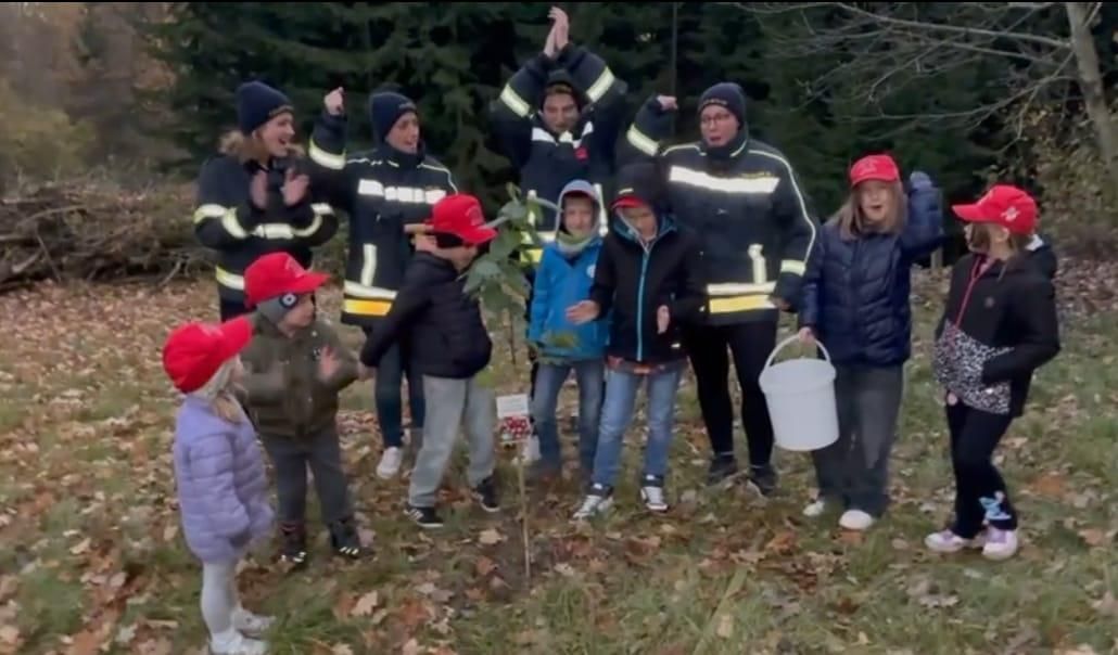 Eine Gruppe von Kindern und Erwachsenen in Winterkleidung versammelt sich um einen Baum, wobei Feuerwehrleute anwesend sind. Sie halten einen Eimer und scheinen an einer Baumpflanzaktion beteiligt zu sein.
