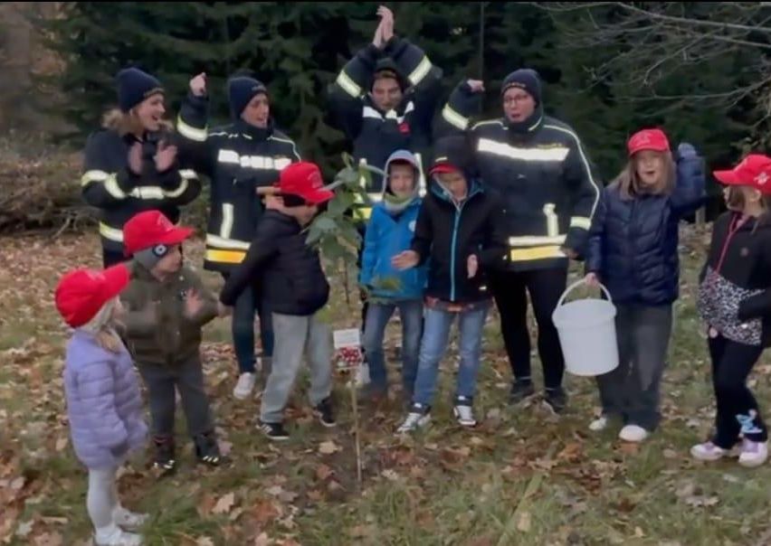 Eine Gruppe von Kindern und Erwachsenen in Winterkleidung versammelt sich um einen Baum, wobei Feuerwehrleute anwesend sind. Sie halten einen Eimer und scheinen an einer Baumpflanzaktion beteiligt zu sein.