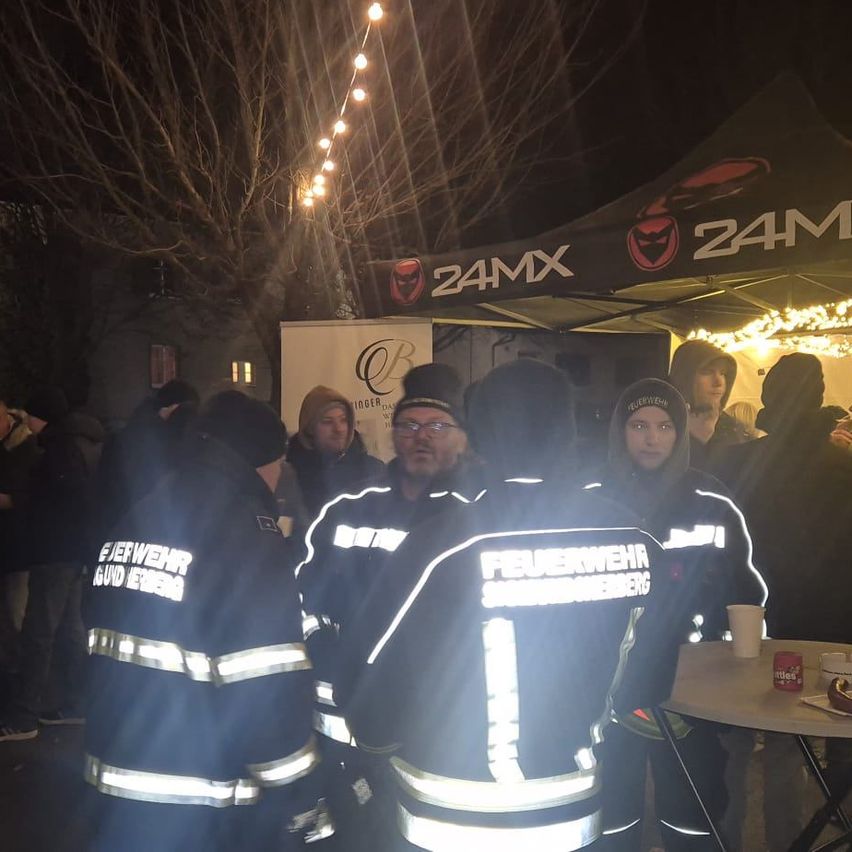 Mehrere Feuerwehrleute in reflektierenden Jacken stehen bei Nacht um einen Tisch herum, unter einem Zelt mit 24MX-Marke.