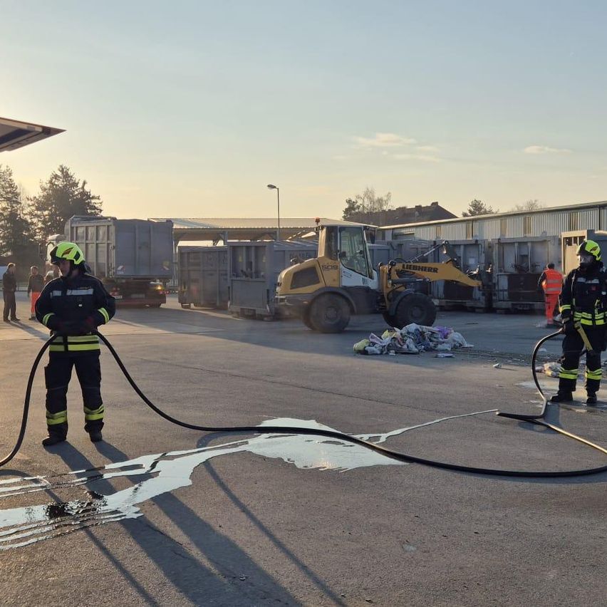 Zwei Feuerwehrleute arbeiten an einer Verschüttung auf der Straße. Ein gelber Baufahrzeug ist in der Nähe geparkt.