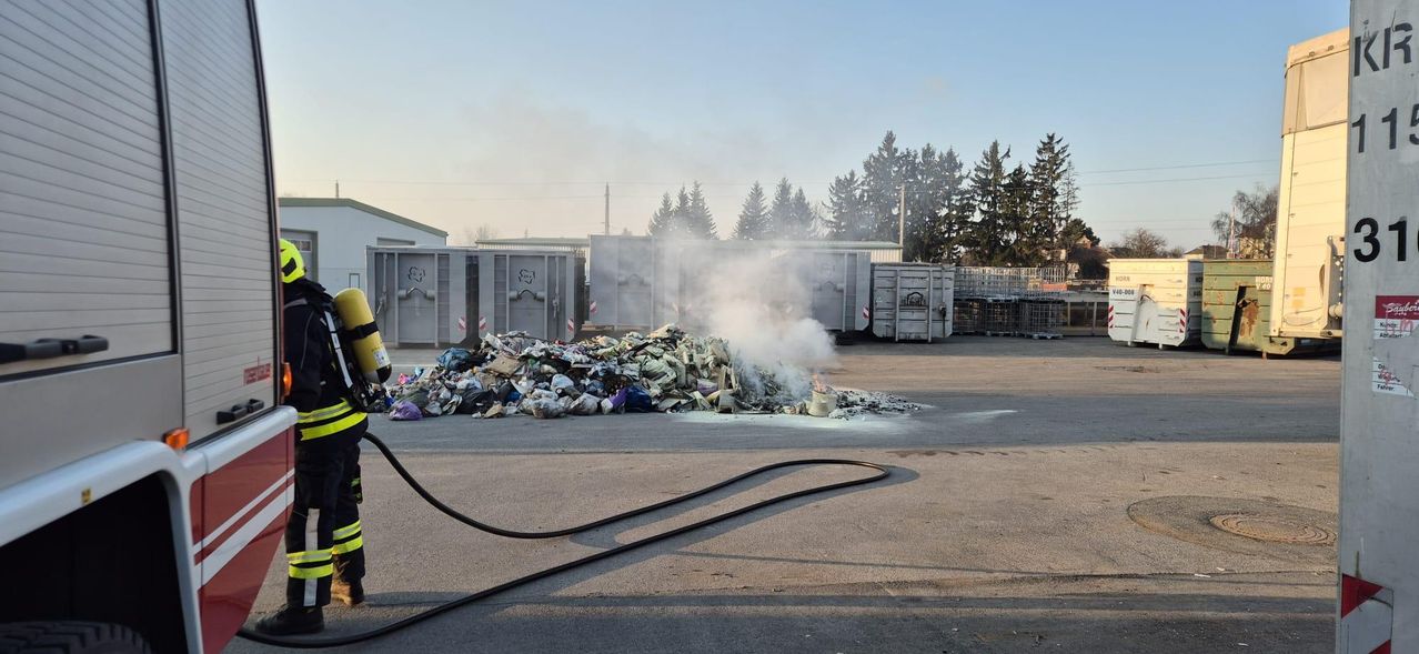 Ein Haufen Abfall brennt draußen mit aufsteigendem Rauch, in der Nähe mehrerer Metallbehälter mit Bäumen im Hintergrund.
