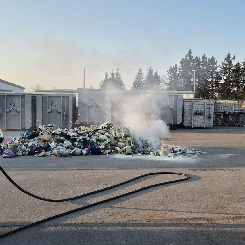 Ein Haufen Abfall brennt draußen mit aufsteigendem Rauch, in der Nähe mehrerer Metallbehälter mit Bäumen im Hintergrund.