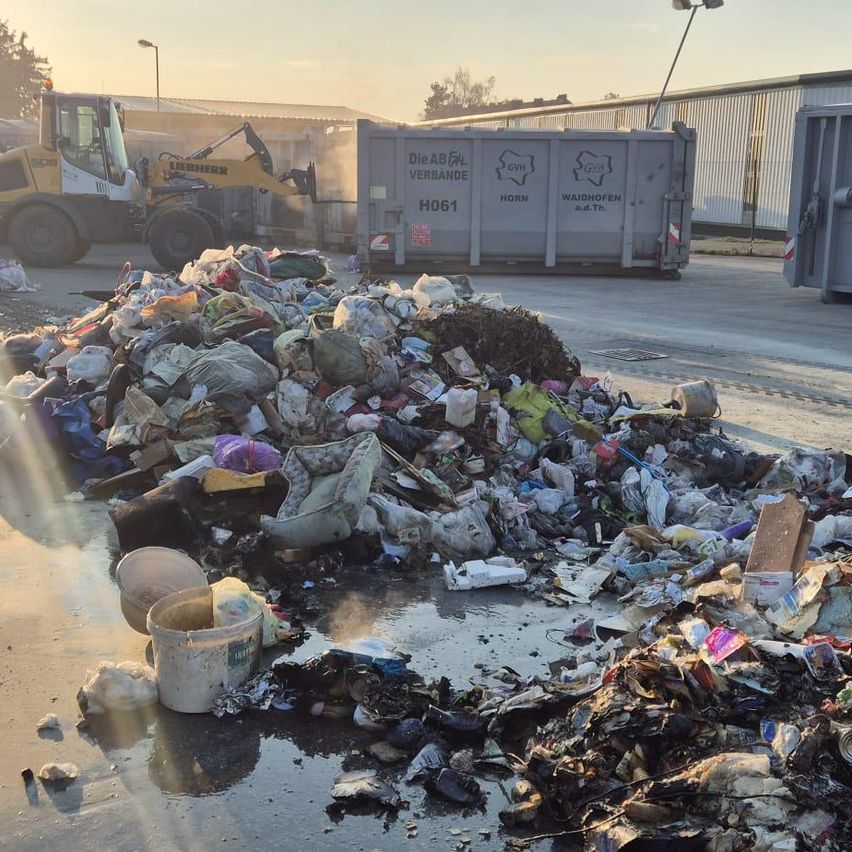 Ein großer Haufen gemischten Mülls und Schutts ist zu sehen, wobei ein gelber Baumaschinenwagen Müll in einen Container entlädt. Der Boden ist nass.