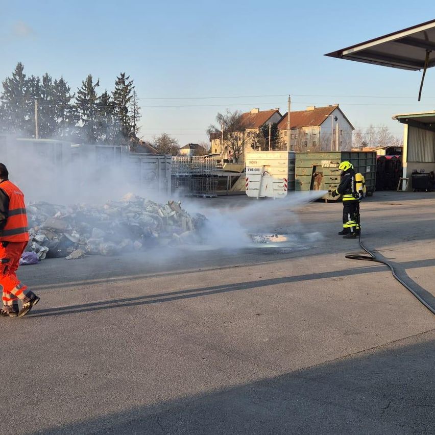 Feuerwehrleute löschen mit einem Schlauch ein Feuer, in der Nähe eines Schutthaufens. Ein Feuerwehrmann steht vorne und sprüht Wasser, während der andere den Schlauch hält.