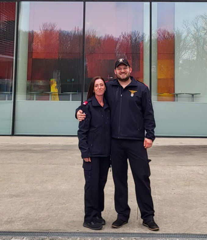 Ein Mann und eine Frau in Uniformen stehen vor einem modernen Gebäude mit Glaswänden und lächeln. Hinter ihnen sind Bäume und Reflexionen sichtbar.