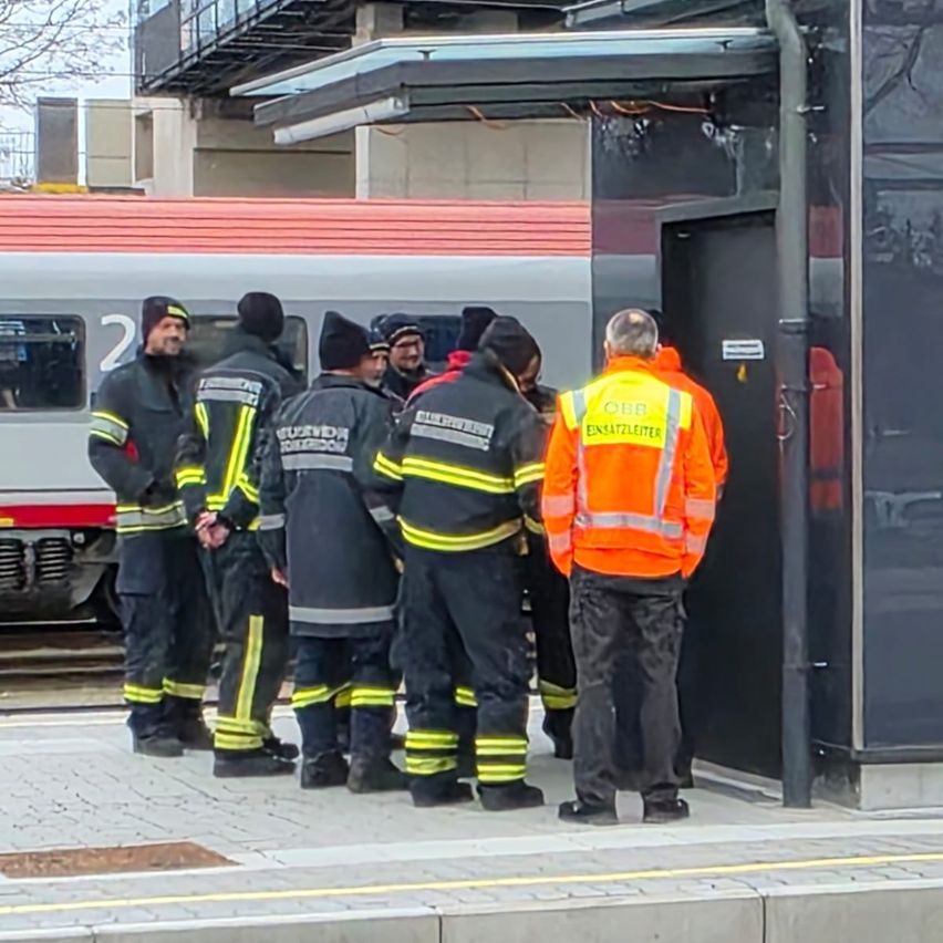Eine Gruppe von Feuerwehrleuten steht auf einem Bahnsteigen der Bahnhofs, einer Mann in einem orangefarbenen Mantel mit reflektierenden Streifen. Ein roter Zug ist hinter ihnen geparkt.