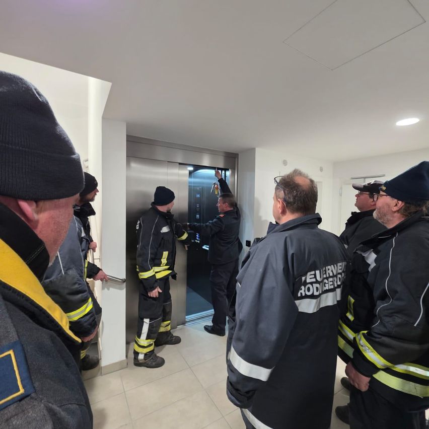 Feuerwehrleute stehen um einen Aufzug herum, wobei ein Mann die Tür offen hält. Sie tragen schwarze Jacken mit reflektierenden Streifen.