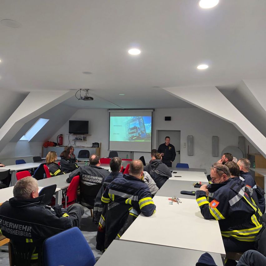 Eine Gruppe Feuerwehrleute in Uniform sitzt an Tischen in einem Klassenzimmer. Ein Mann steht vor einer Projektionsleinwand und hält eine Präsentation.
