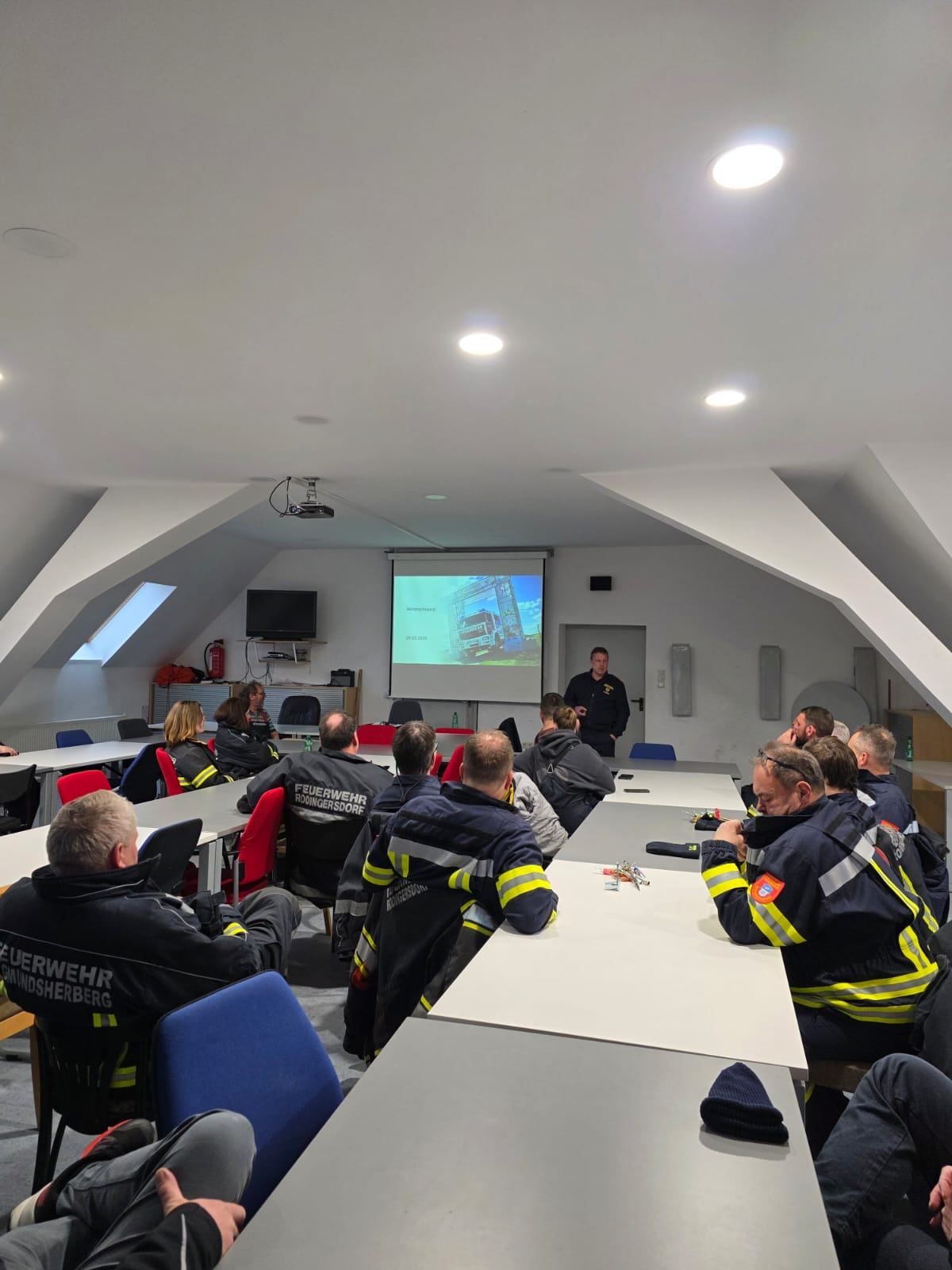 Eine Gruppe Feuerwehrleute in Uniform sitzt an Tischen in einem Klassenzimmer. Ein Mann steht vor einer Projektionsleinwand und hält eine Präsentation.