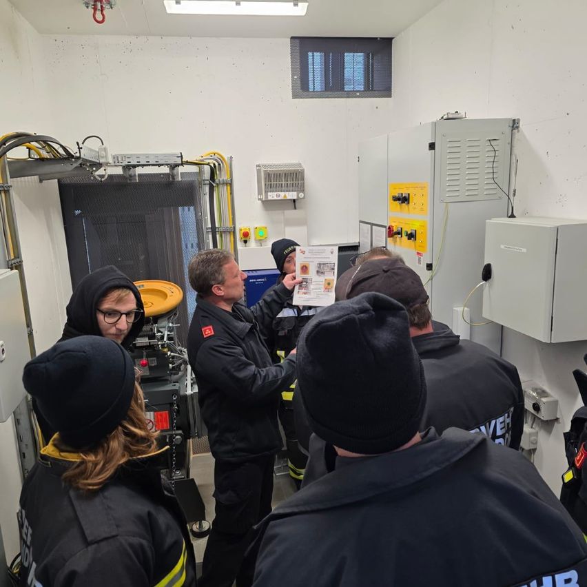 Eine Gruppe von Personen in schwarzen Uniformen und Mützen versammelt sich in einem Raum, möglicherweise einer Schulungseinrichtung. Ein Mann in der Mitte hält ein Papier und erklärt den Anwesenden etwas.