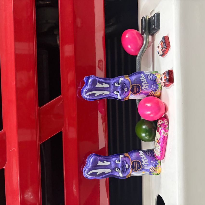 Zwei lila Schokoladenhasenfiguren hängen an einer roten Tür neben pinken und grünen Eiern. Ein kleiner roter Haken hält sie an Ort und Stelle.