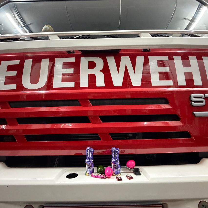 Ein roter Feuerwehrwagen mit 'FEUERWEHR' in weiß auf der Vorderseite geschrieben. Mehrere Schokoladenfiguren sind auf dem Stoßfänger zu sehen, darunter ein rosa Osterhase, grüne Schokoladeneier und andere Osterleckerlichkeiten.