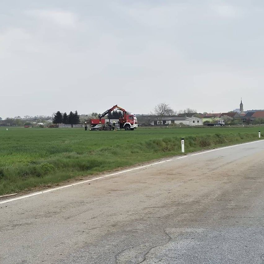 Ein roter Kran steht auf einem Feld, mit Menschen, die darum stehen. Eine Straße ist auf der rechten Seite mit weißen Markierungen. Ein paar Gebäude sind in der Ferne.