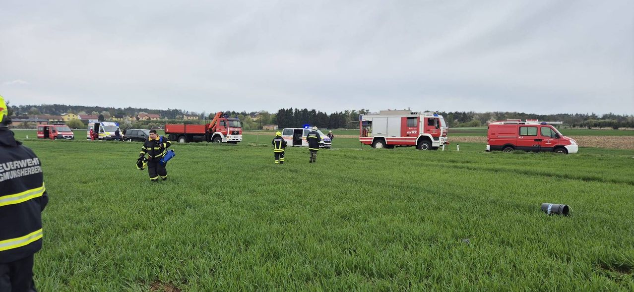 Rettungsfahrzeuge sind auf einer Wiese geparkt. Feuerwehrleute stehen mit Helmen auf dem Feld. Ein Van und ein Lkw sind in der Nähe geparkt. Bäume und Gebäude sind im Hintergrund sichtbar.