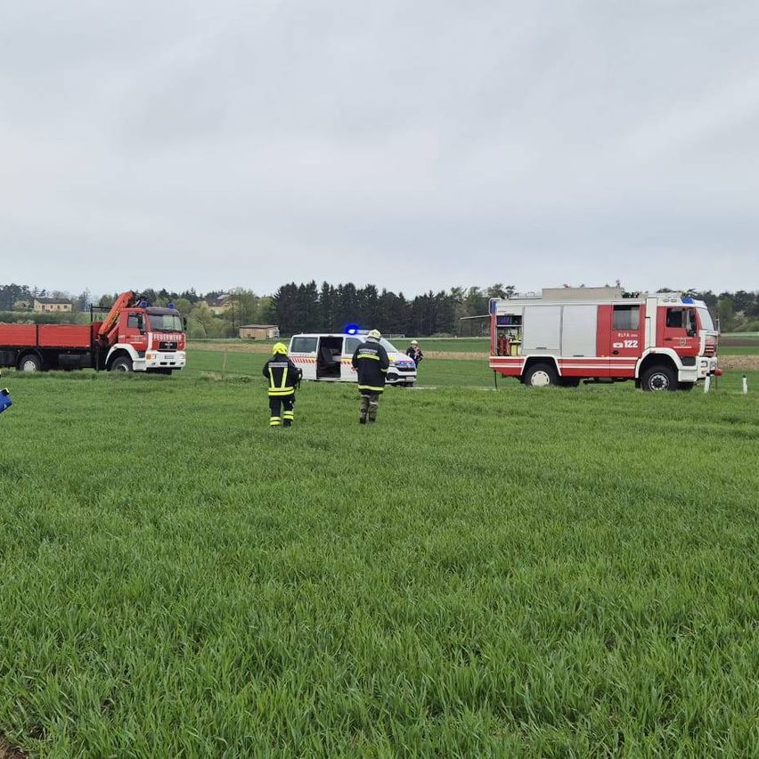 Rettungsfahrzeuge sind auf einer Wiese geparkt. Feuerwehrleute stehen mit Helmen auf dem Feld. Ein Van und ein Lkw sind in der Nähe geparkt. Bäume und Gebäude sind im Hintergrund sichtbar.