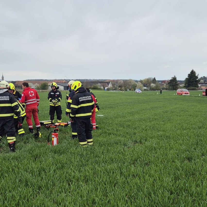 Eine Gruppe von Notfallhelfern in Uniform steht in einem Grasfeld mit einem Feuerlöscher. In der Ferne sind Fahrzeuge geparkt, und Gebäude sind sichtbar.