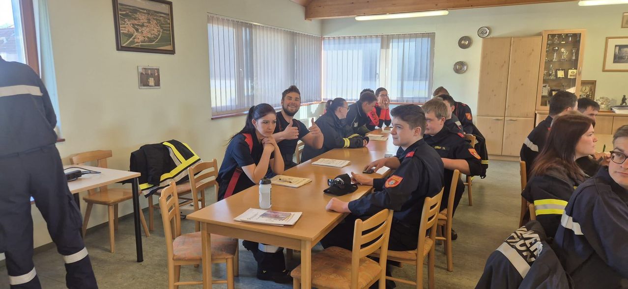 Eine Gruppe von Feuerwehrleuten, einige in Uniform, sind um einen Tisch versammelt. Sie lächeln, lachen und machen Gesten. Sie scheinen eine Besprechung oder Schulung zu haben.