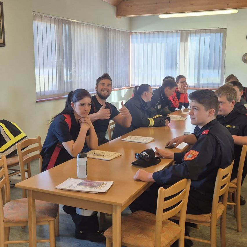 Eine Gruppe von Feuerwehrleuten, einige in Uniform, sind um einen Tisch versammelt. Sie lächeln, lachen und machen Gesten. Sie scheinen eine Besprechung oder Schulung zu haben.