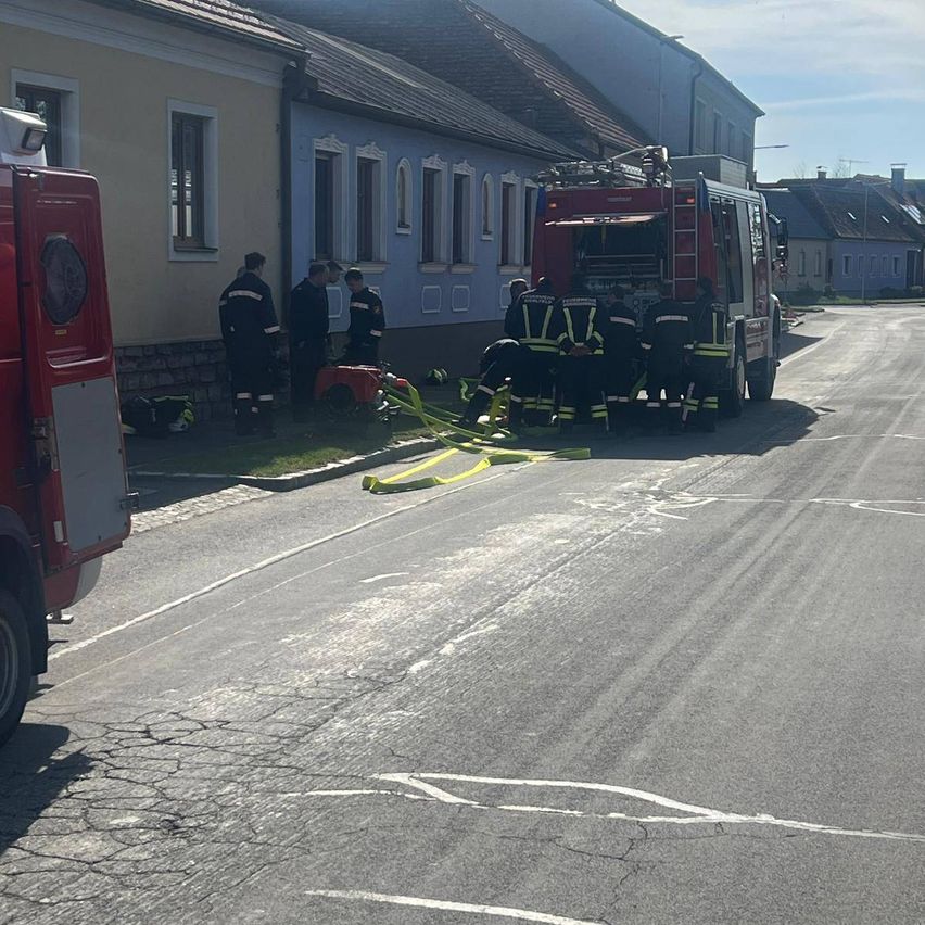 Feuerwehrleute versammeln sich auf einer Straße in der Nähe eines Feuerwehrwagens. Ein Feuerlöscher liegt auf dem Boden. Die Feuerwehrleute scheinen etwas zu besprechen.