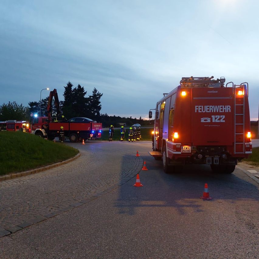 Ein Feuerwehrauto mit eingeschalteten Lichtern steht am Straßenrand, mit mehreren Verkehrskegeln und Personen in Helmen.