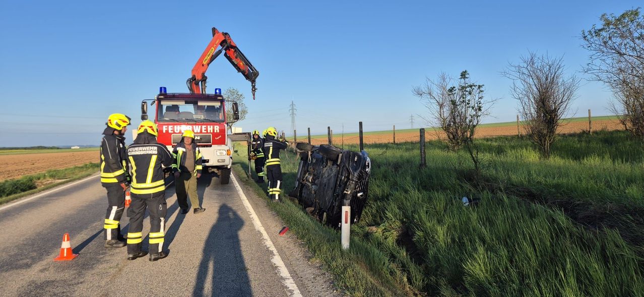Ein Feuerwehrauto mit einem Kran hebt ein umgekipptes Auto von der Straße. Rettungskräfte stehen mit Helmen um das Auto herum.