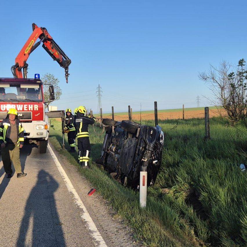Ein Feuerwehrauto mit einem Kran hebt ein umgekipptes Auto von der Straße. Rettungskräfte stehen mit Helmen um das Auto herum.