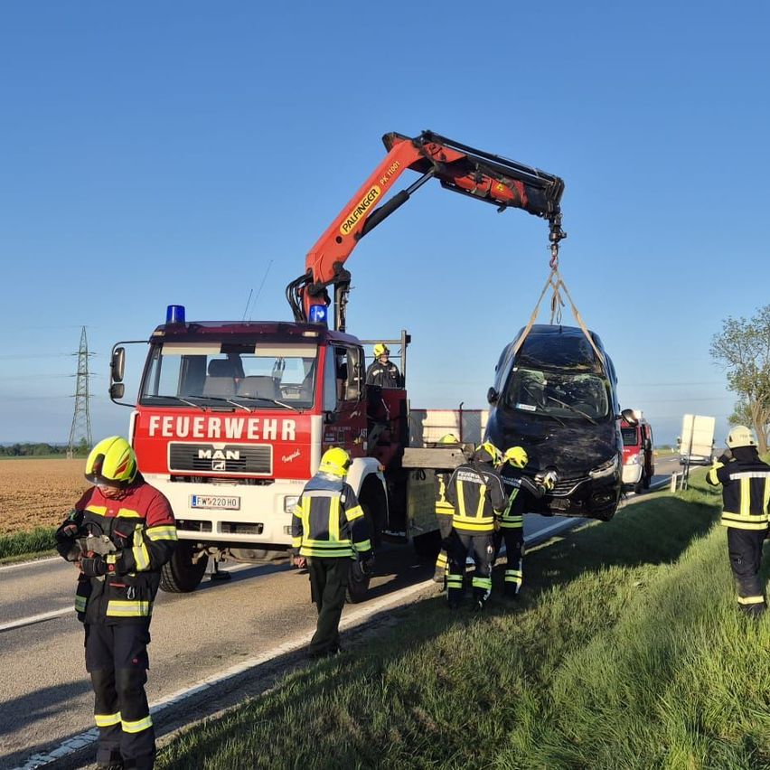 Ein Feuerwehrwagen hebt ein verunglücktes Auto von der Straße. Mehrere Feuerwehrleute in Schutzausrüstung helfen. Das Auto ist beschädigt mit Rauch und Staub.