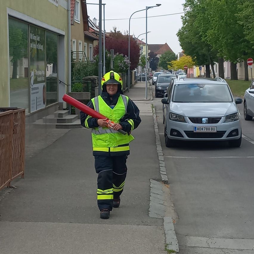 Ein Feuerwehrmann in voller Ausrüstung geht mit einem roten Stab die Straße entlang, neben geparkten Autos.