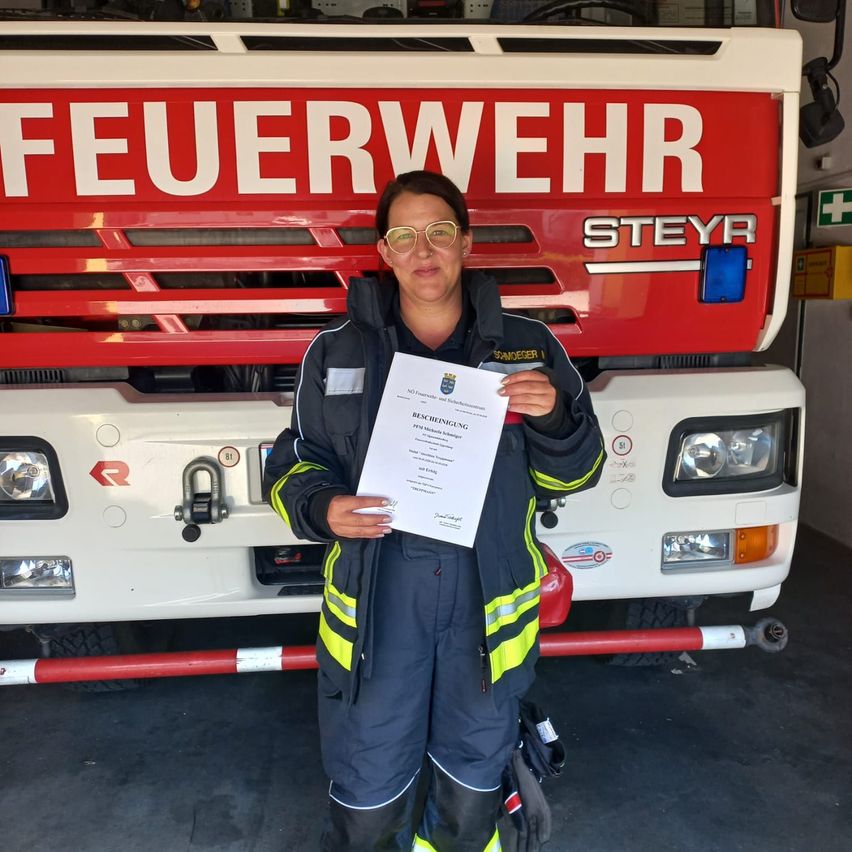 Eine Frau in Feuerwehruniform steht vor einem Feuerwehrwagen und hält ein Zertifikat in der Hand. Der Wagen hat das Wort 'Feuerwehr' auf seiner Vorderseite.
