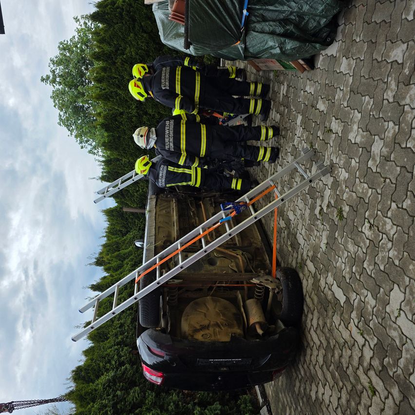 Drei Feuerwehrleute in gelben und schwarzen Uniformen stehen mit Leitern auf einem Dach, einer scheint die Unterseite des Autos zu inspizieren.