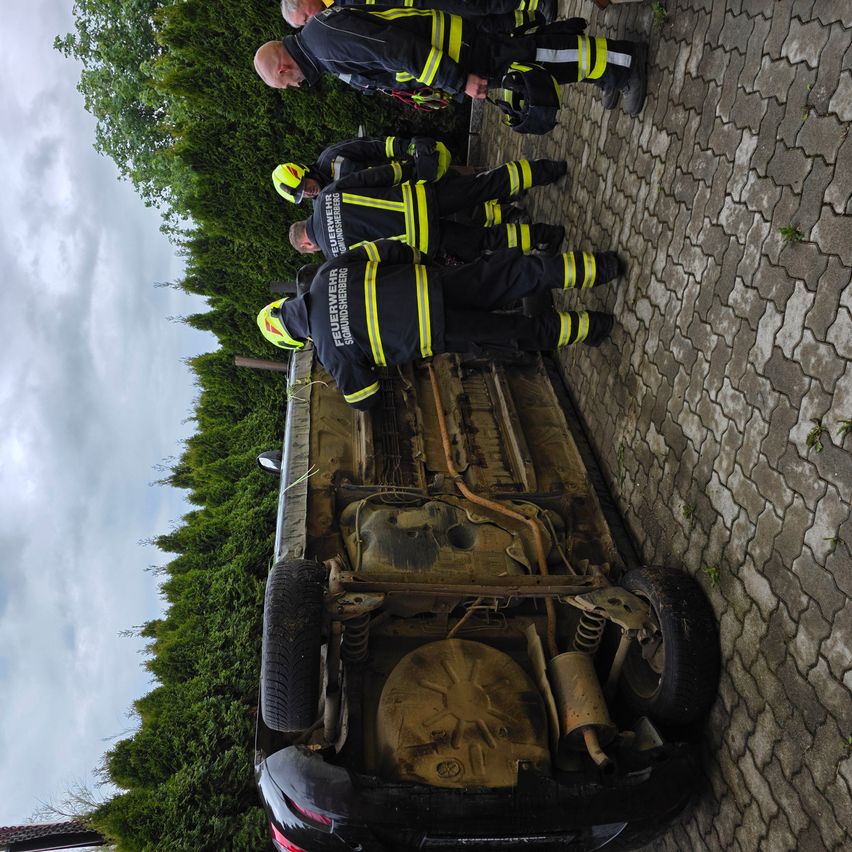 Feuerwehrleute stehen neben einem umgekippten Auto auf einer Ziegelstraße, wobei eine Person das Fahrzeug untersucht.