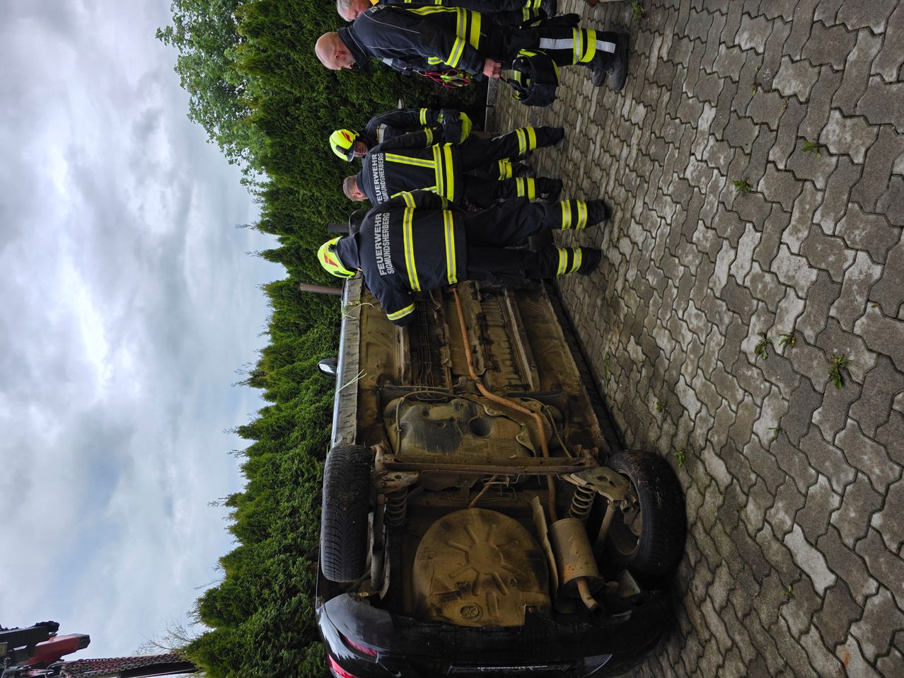 Feuerwehrleute stehen neben einem umgekippten Auto auf einer Ziegelstraße, wobei eine Person das Fahrzeug untersucht.