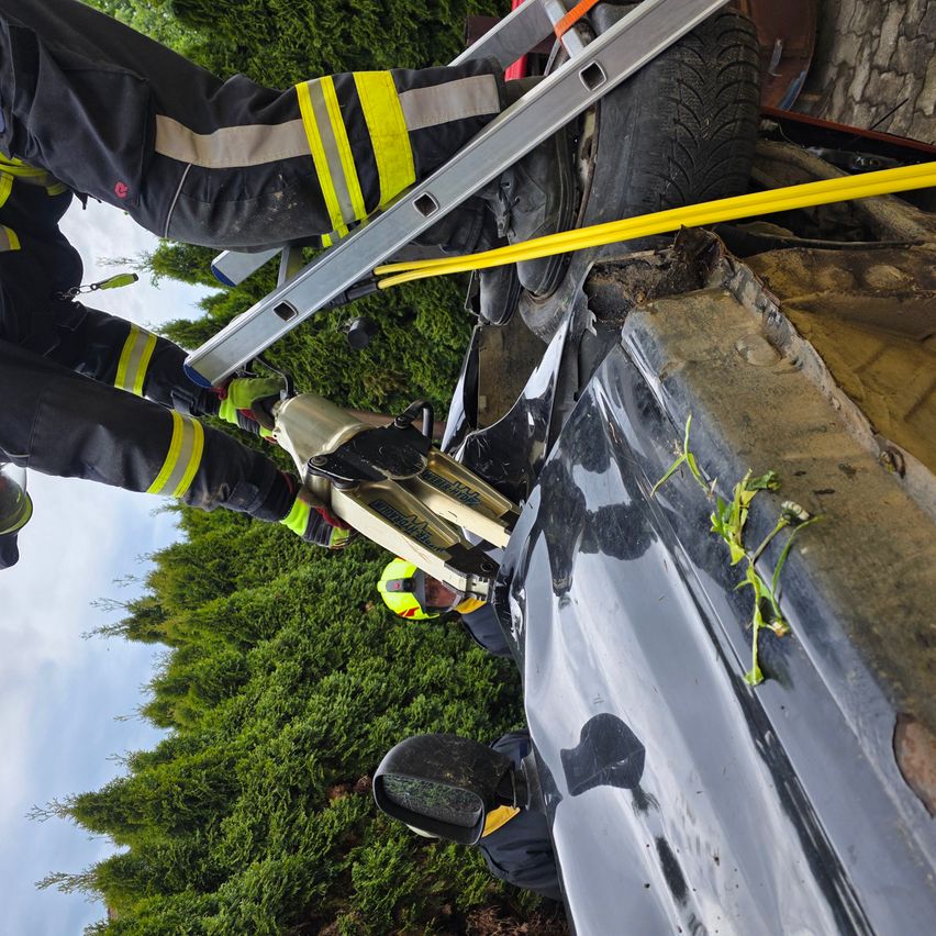 Feuerwehrleute retten eine Person aus einem beschädigten Auto mit einer Leiter. Das Auto ist kaputt und die Rettungsaktion findet im Freien statt.