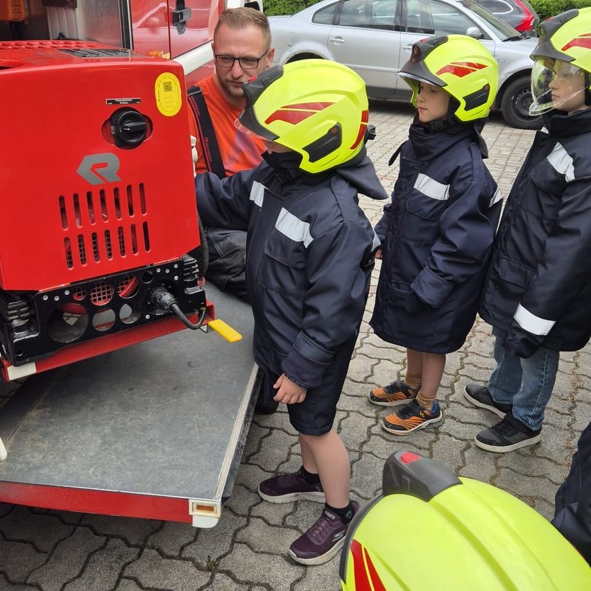 Ein Mann und vier Kinder in Feuerwehruniformen untersuchen ein Feuerwehrfahrzeug. Die Kinder tragen gelbe Helme.