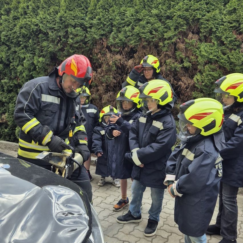 Ein Feuerwehrmann zeigt Kindern in Helmen, wie man einen Feuerlöscher in der Nähe eines Autos benutzt, mit Hecken im Hintergrund.