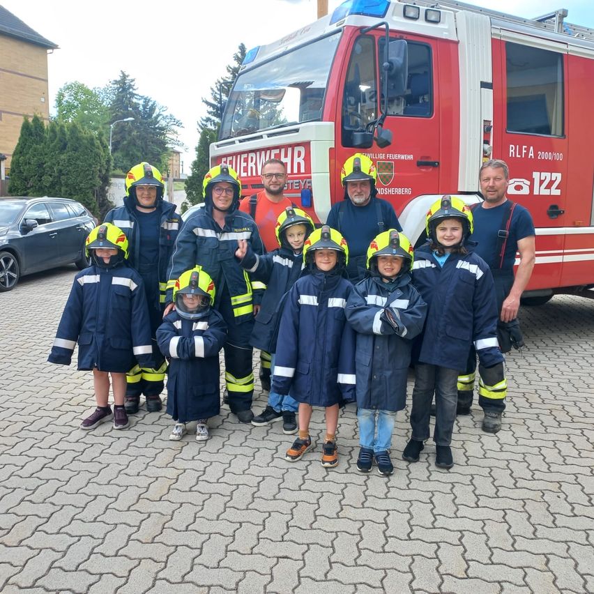 Eine Gruppe Feuerwehrleute und Kinder in Schutzkleidung stehen vor einem roten Feuerwehrwagen. Dahinter sind ein Gebäude und ein geparktes Auto zu sehen.