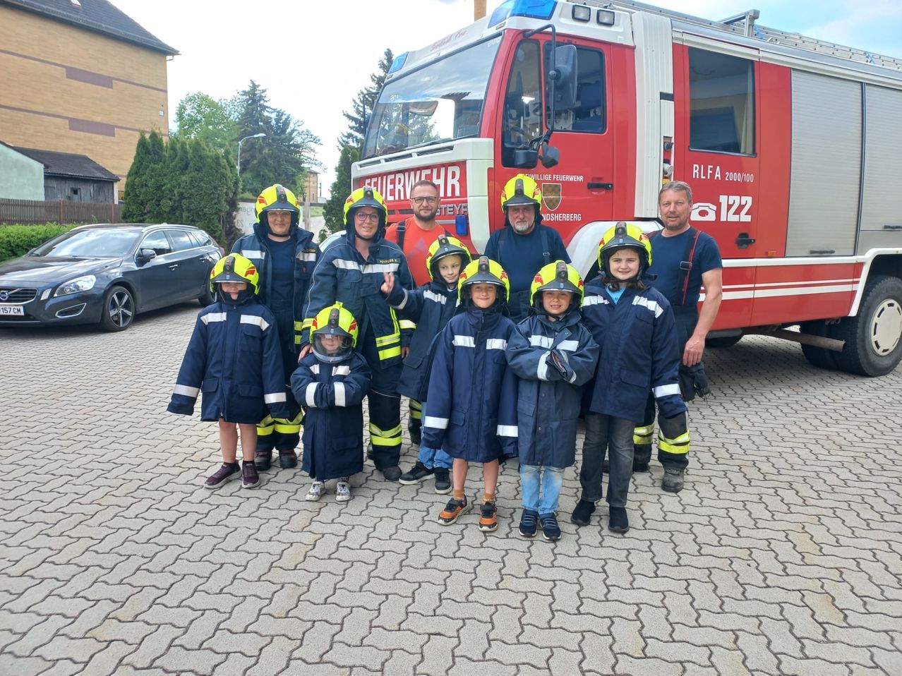 Eine Gruppe Feuerwehrleute und Kinder in Schutzkleidung stehen vor einem roten Feuerwehrwagen. Dahinter sind ein Gebäude und ein geparktes Auto zu sehen.