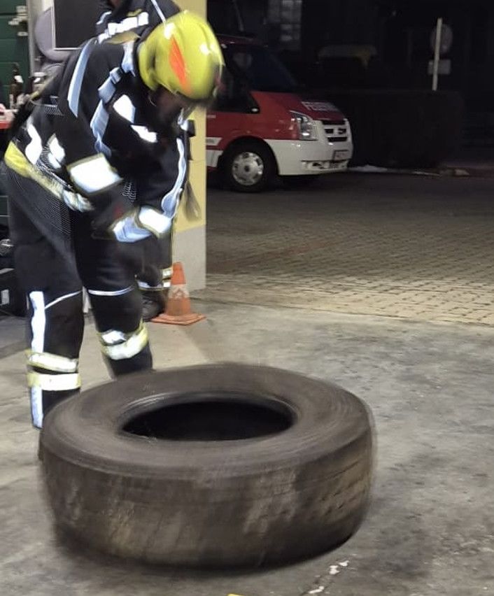 Ein Feuerwehrmann übt mit einem Werkzeug an einem Reifen auf einem Parkplatz. Ein Van ist hinter ihm geparkt.