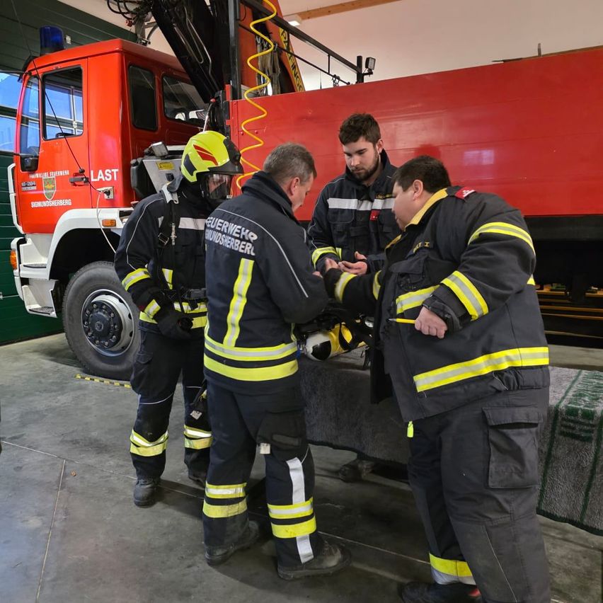 Vier Feuerwehrleute in voller Ausrüstung stehen um ein Feuerwehrauto herum. Einer hält einen Helm, während ein anderer darauf zeigt. Der Ort ist eine Feuerwache.