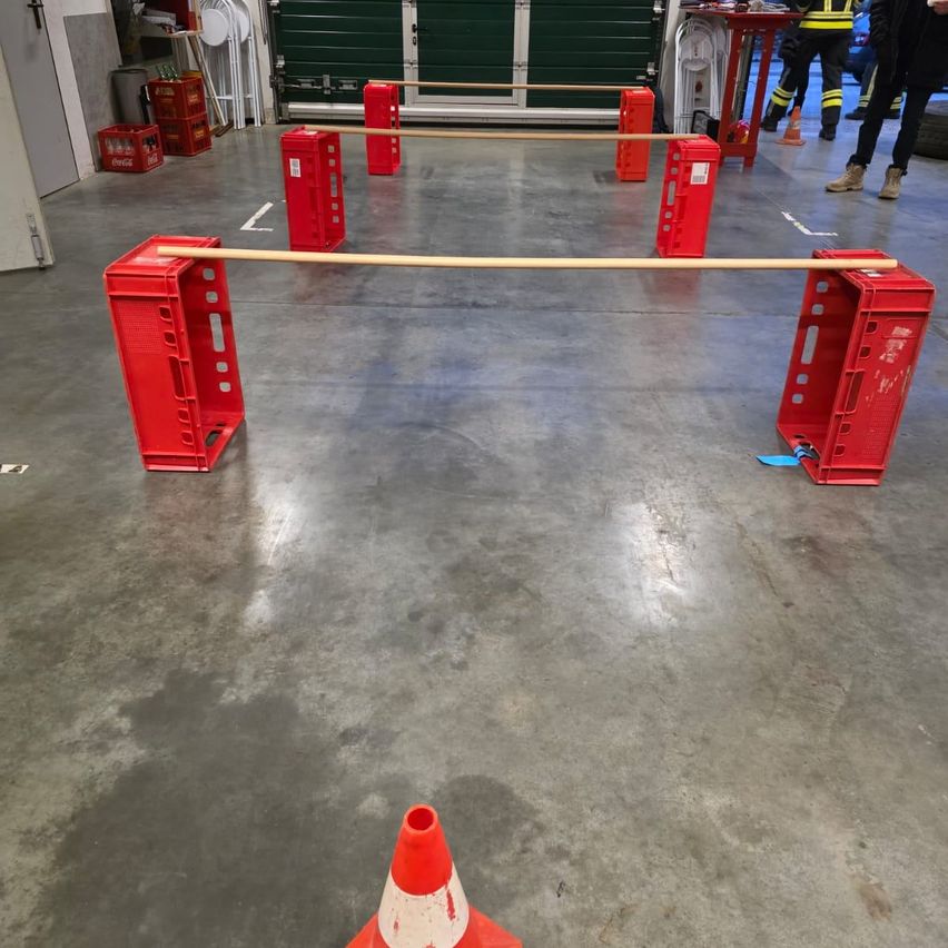 Ein Lager mit roten Kisten, die eine Barriere bilden, einem langen Holzstab und einem orangefarbenen Verkehrskegel. Im Hintergrund sind Personen zu sehen.